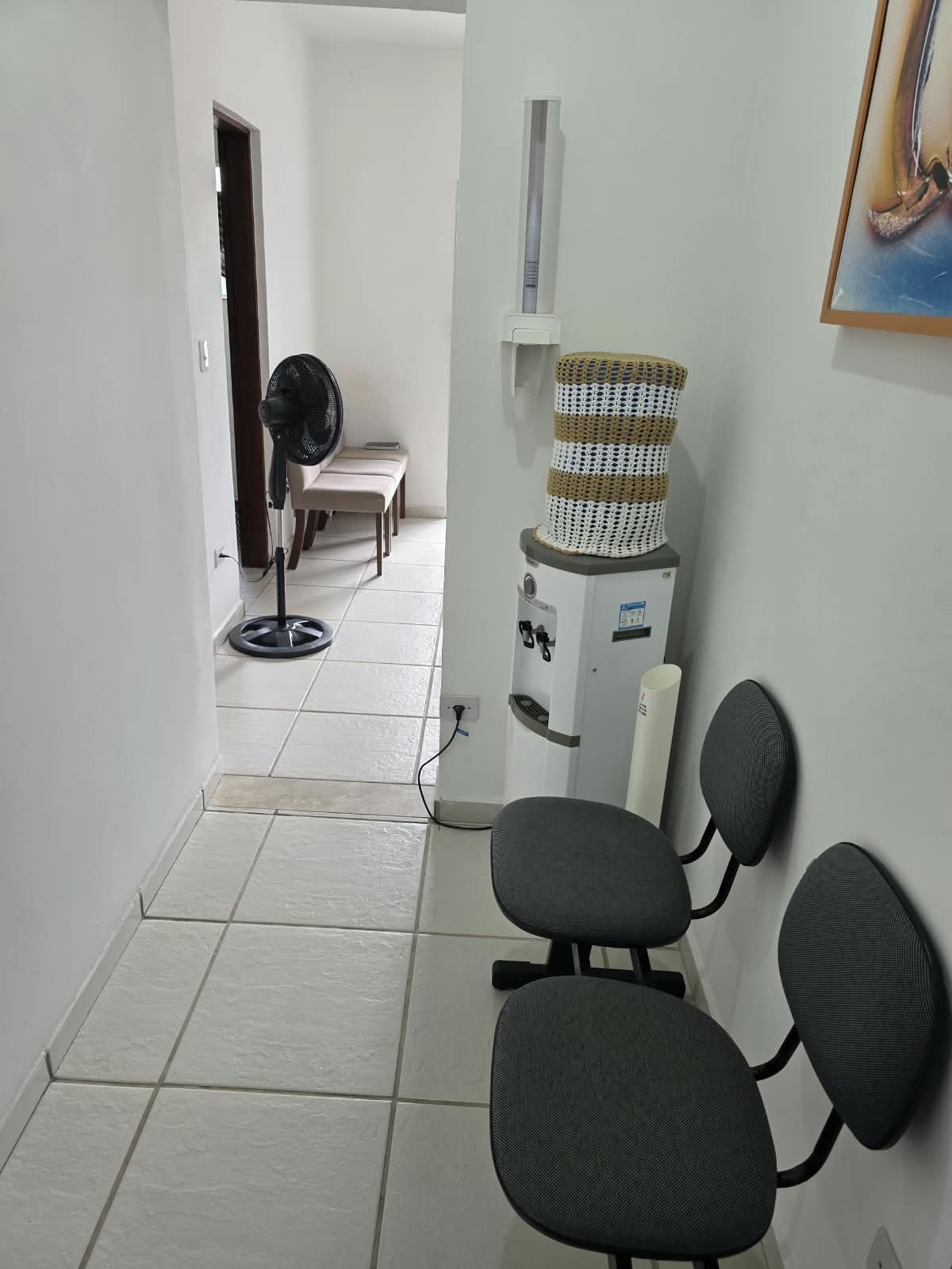 Corredor interno com cadeiras, ventilador e bebedouro.
