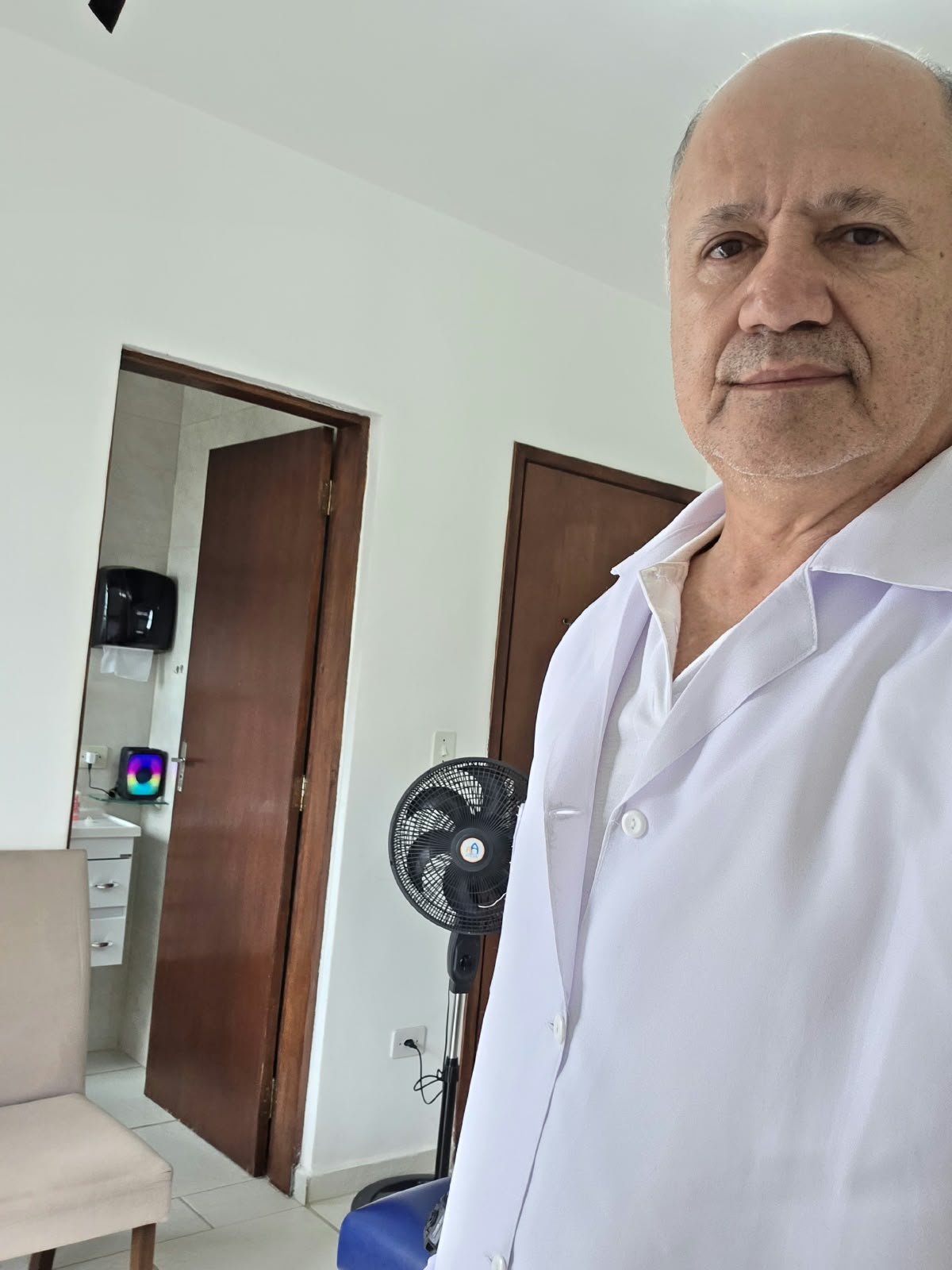 Profissional de jaleco no consultório, com maca e uma sala ao fundo.
